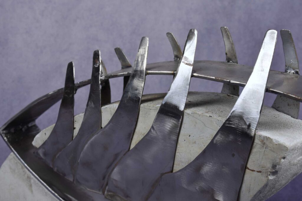 Close-up side view: steel and concrete sculpture; steel ribs overlapping and growing larger from front to back hold a v-shaped concrete block. All suspended from steel bar shaped like a teardrop facing forward.