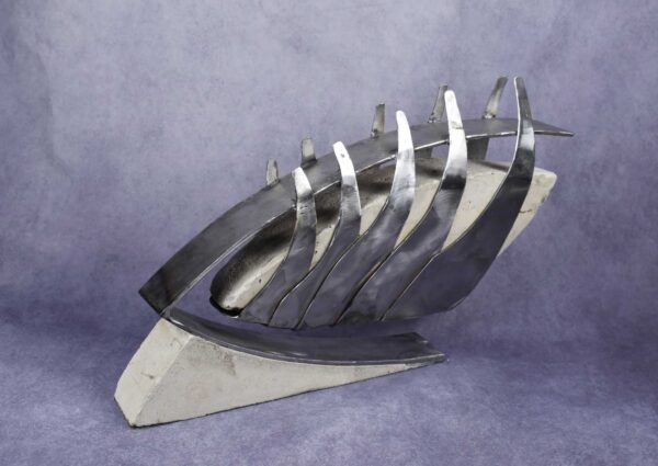 Three quarter view: steel and concrete sculpture; steel ribs overlapping and growing larger from front to back hold a v-shaped concrete block. All suspended from steel bar shaped like a teardrop facing forward.