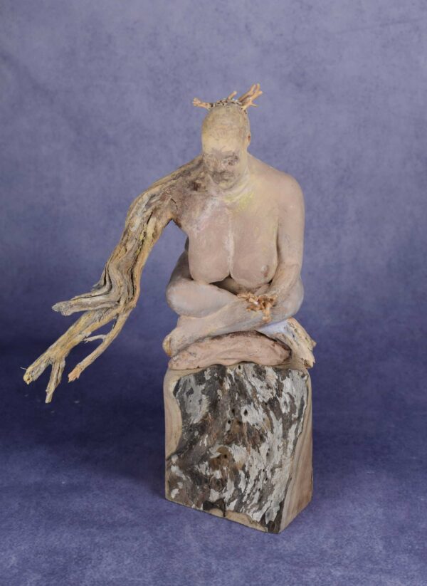 Front view: clay sculpture of seated woman with a branch for an arm and coral protruding from her head like a crown.