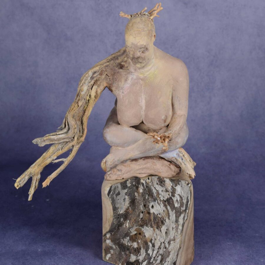 Front view: clay sculpture of seated woman with a branch for an arm and coral protruding from her head like a crown.