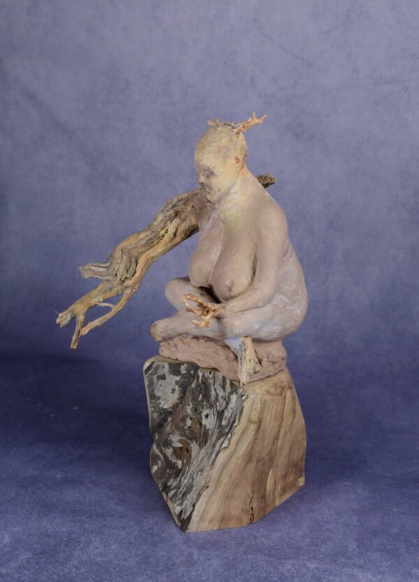 Left side view: clay sculpture of seated woman with a branch for an arm and coral protruding from her head like a crown.