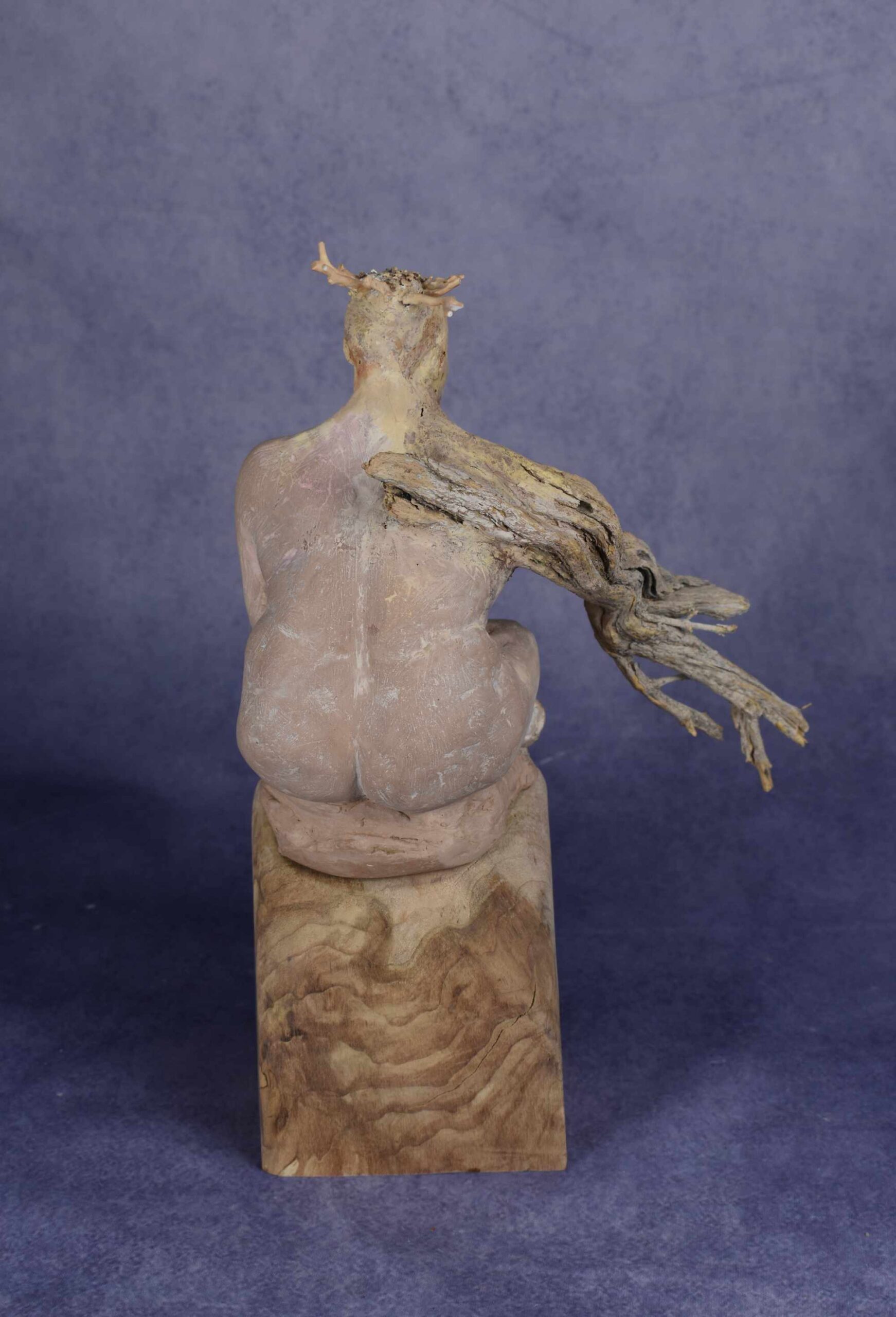 Back view: clay sculpture of seated woman with a branch for an arm and coral protruding from her head like a crown.